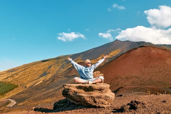 Happy tourist woman enjoying freedom while doing yoga on big stone at panoramic view of colorful summits of active volcano Etna, Tallest volcano in Continental Europe, Sicily, Italy.